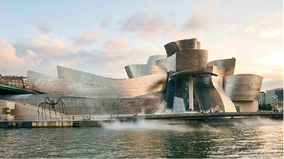 A large, silver, curvilinear museum sits by the waterfront in Bilbao, Spain.  